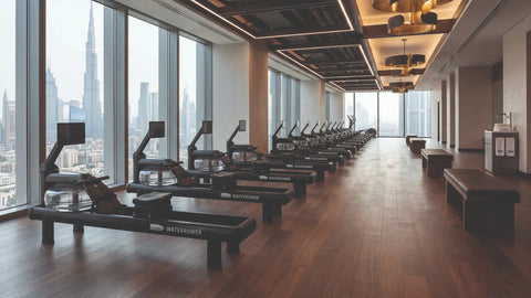 A row of WaterRower rowing machines arranged in a modern fitness gym with large windows overlooking a city skyline, featuring dark wood floors and ceiling fixtures.