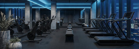 A modern gym interior with rows of NOHRD Citius treadmills and Bikes in a dark, high-end fitness space with blue ambient lighting and large windows overlooking a city skyline.