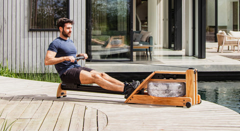 A man using a wooden WaterRower Hybrid rowing machine on an outdoor deck in front of a modern wooden house.