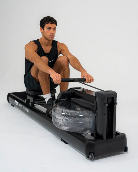 A man wearing a black Nike tank top and shorts is rowing on a black WaterRower Metallic against a clean white studio background.